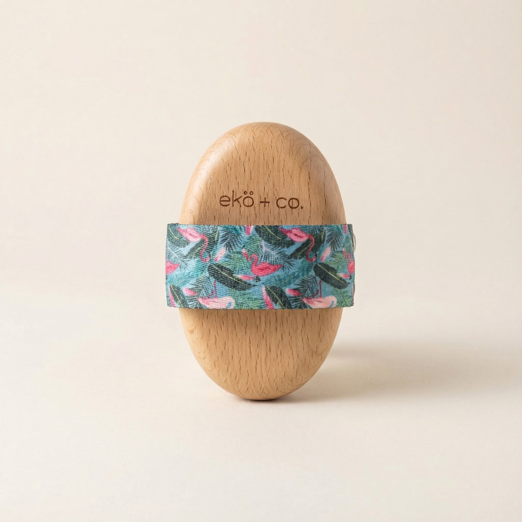 beechwood dry body brush standing upright with tropical flamingo-patterned cotton strap wrapped around middle against neutral beige background