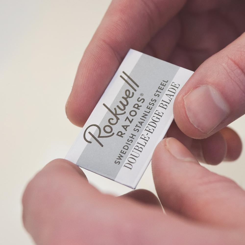 Hand holding individual Rockwell Razors blade wrapper showing Swedish stainless steel double-edge blade ready for installation in safety razor.