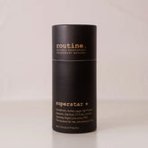 Routine Superstar deodorant in a matte-black cylindrical paper tube with gold “superstar” text, standing on a light beige surface against a matching beige backdrop.