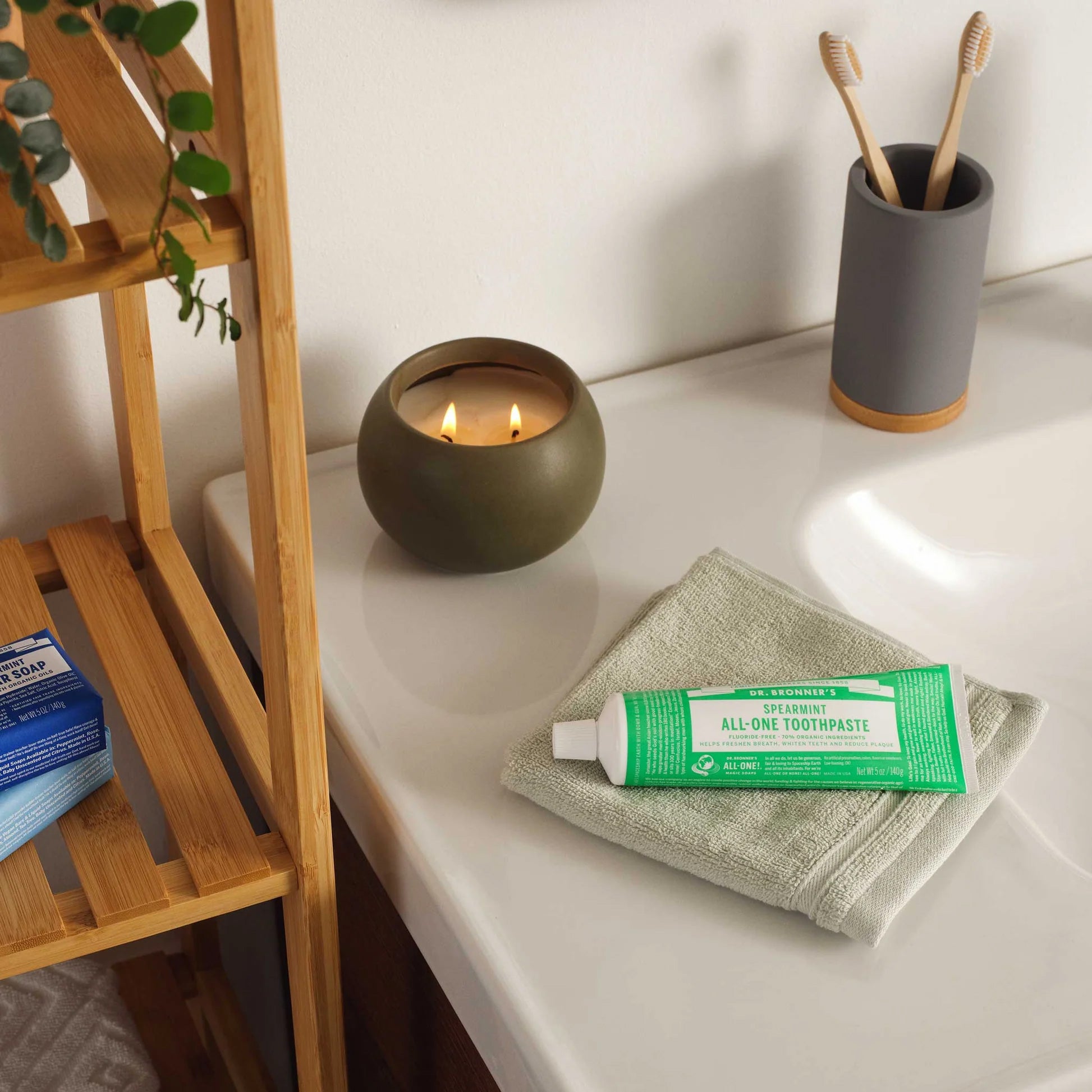 Zen bathroom setting with Dr. Bronner's Spearmint toothpaste on natural linen cloth beside bamboo toothbrushes in grey holder and olive candle