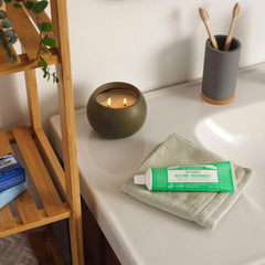 Zen bathroom setting with Dr. Bronner's Spearmint toothpaste on natural linen cloth beside bamboo toothbrushes in grey holder and olive candle