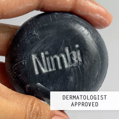 Hand holding wet Nimbi charcoal shave puck with embossed logo, marked Dermatologist Approved