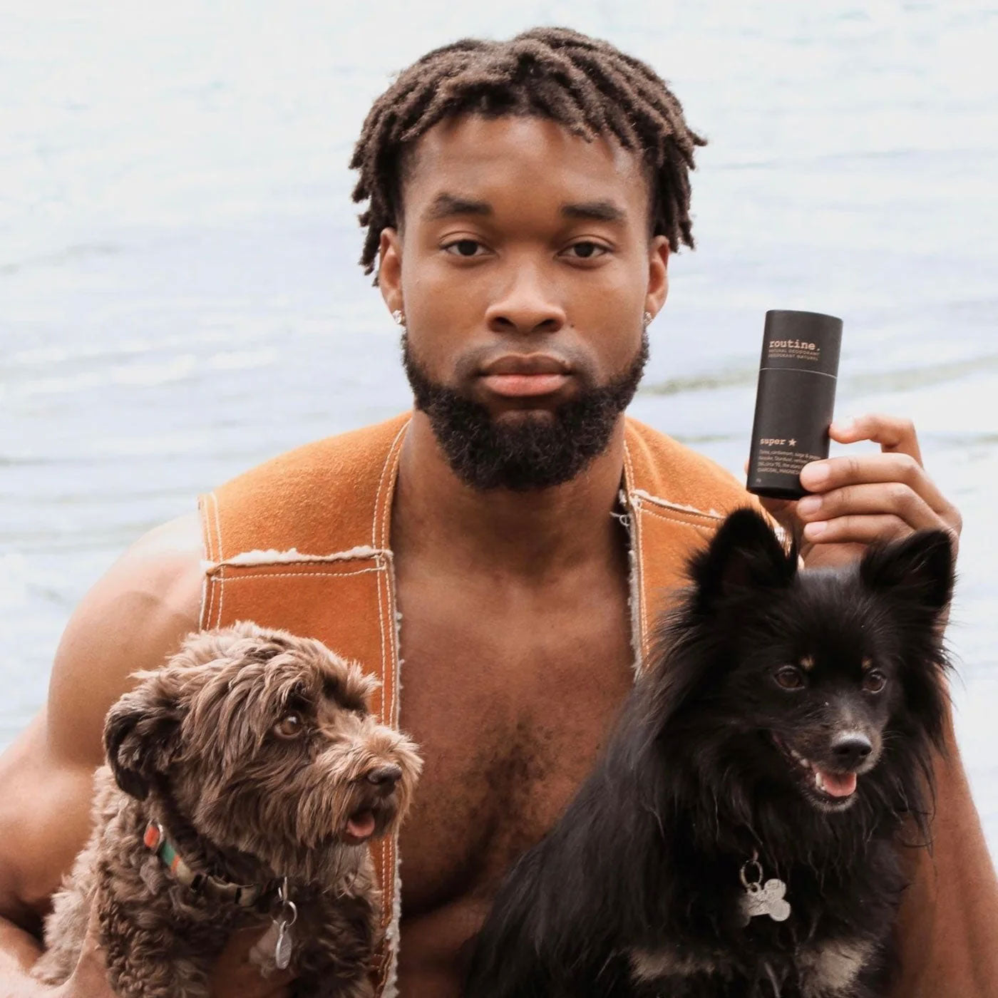 A man holding the Routine Superstar deodorant stick by a lake, flanked by two small dogs, showcasing the product in an outdoor setting.