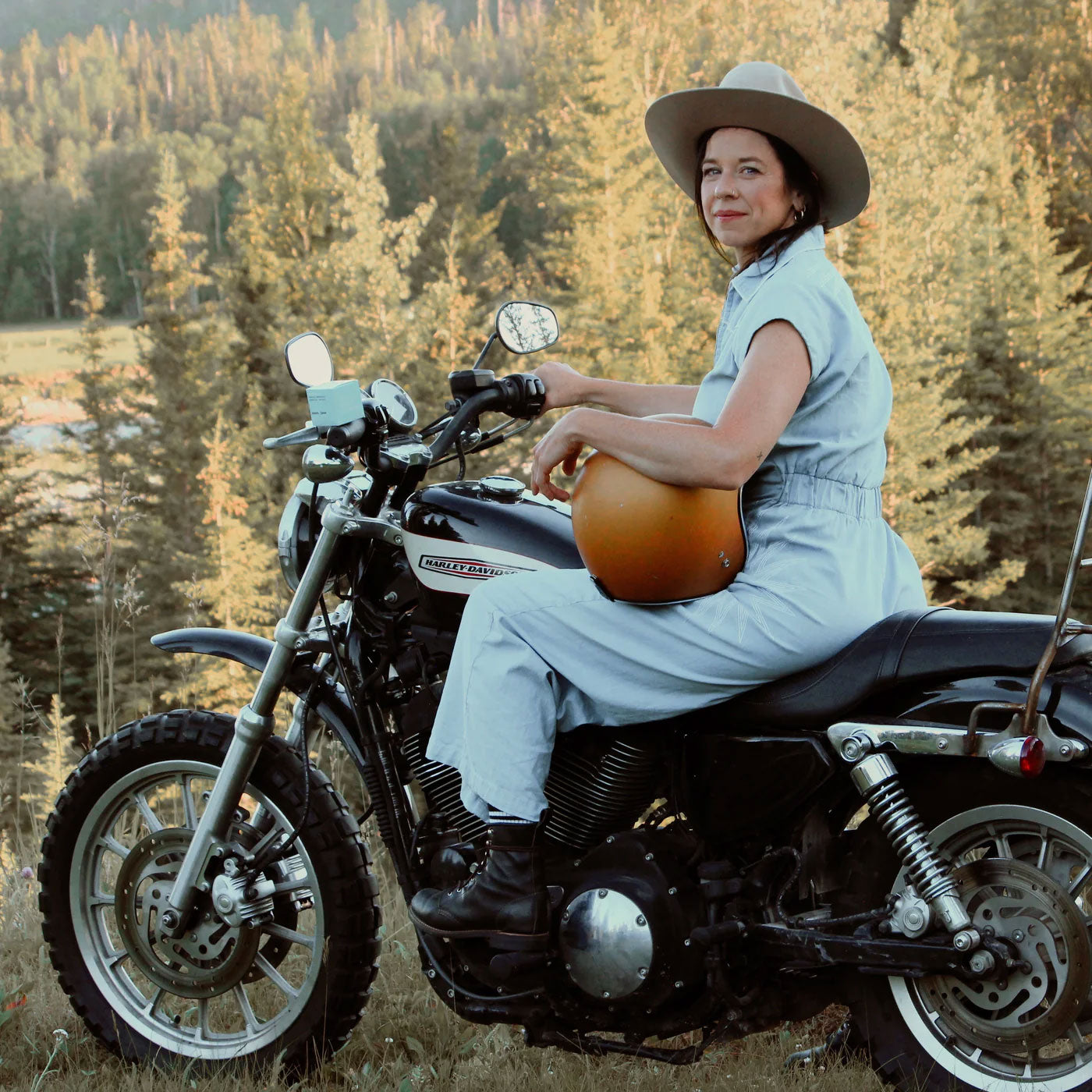 Sweet Jane deodorant brand storytelling - woman in denim overalls and wide-brimmed hat sitting on Harley Davidson motorcycle in golden hour countryside - embodying the grown-up Jane character living her best life in upstate Alberta, Canada