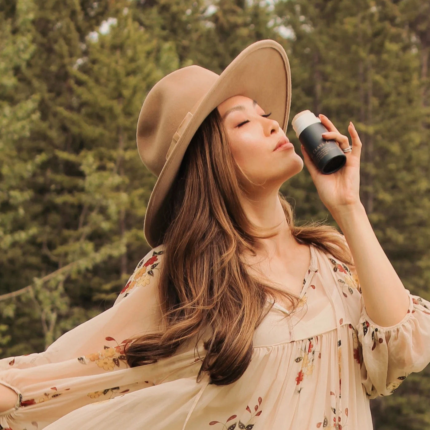 A woman standing in front of evergreen trees, wearing a wide-brimmed tan hat and a cream floral blouse, holding a navy-blue and white Routine The Class deodorant stick up to her nose with her eyes closed, as if savoring its scent.
