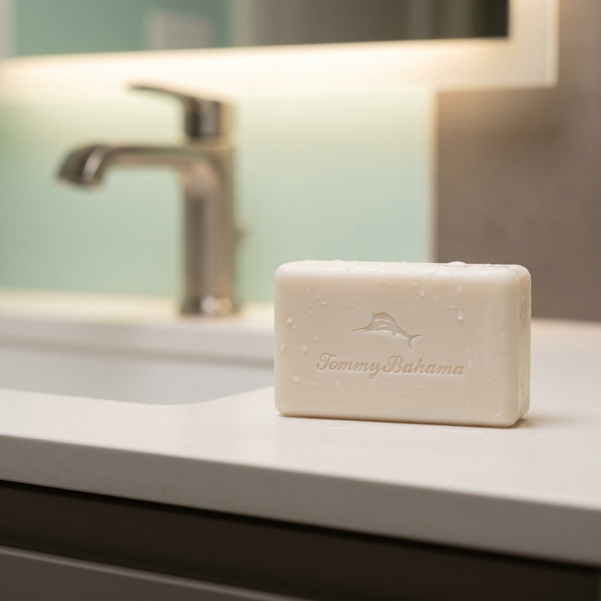Unwrapped Tommy Bahama bar soap with embossed marlin logo on a white bathroom counter beside a chrome faucet, lightly beaded with water droplets